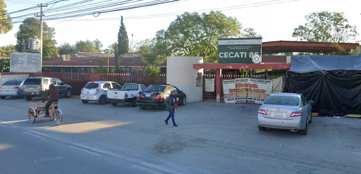 Fotografía en la que se muestra la entrada del plantel, conformada por una reja de color rojo que rodea las instalaciones. Encima de una de las rejas hay una placa de color verde que en letras blancas dice CECATI 85, esta reja da acceso a las instalaciones. 